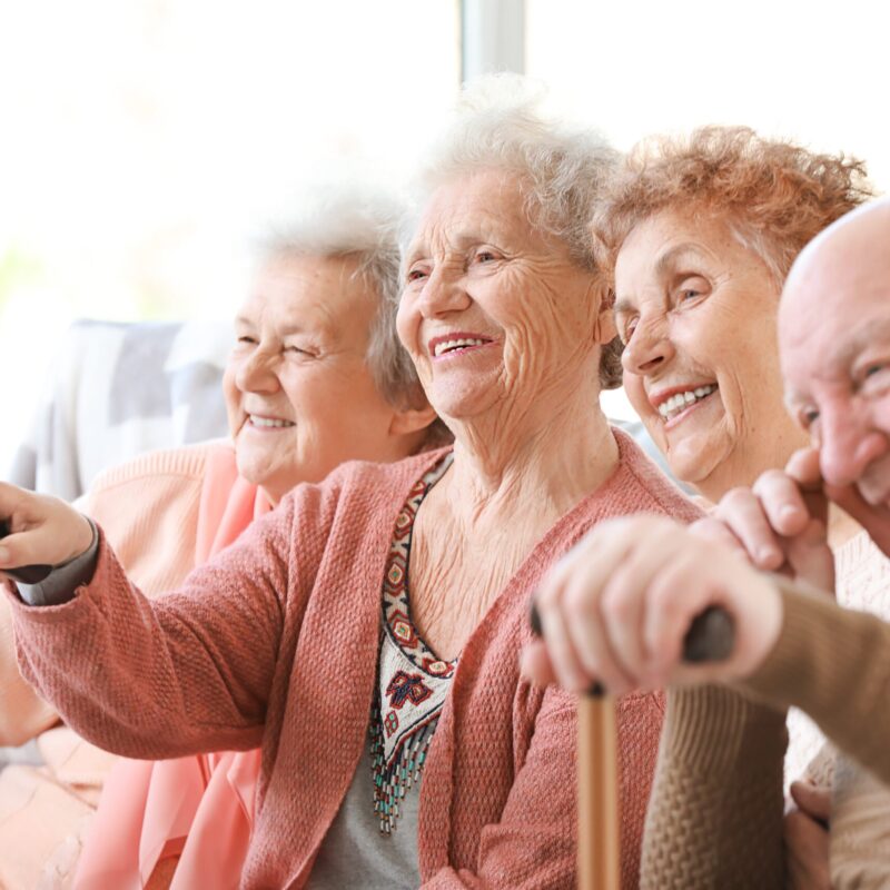 Groupe de 4 seniors assis sur le canapé avec des télécommandes dans les mains regardant la télé et souriant
