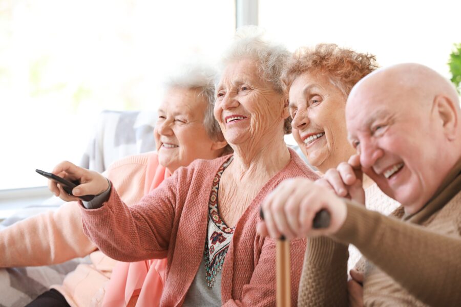 Groupe de 4 seniors assis sur le canapé avec des télécommandes dans les mains regardant la télé et souriant