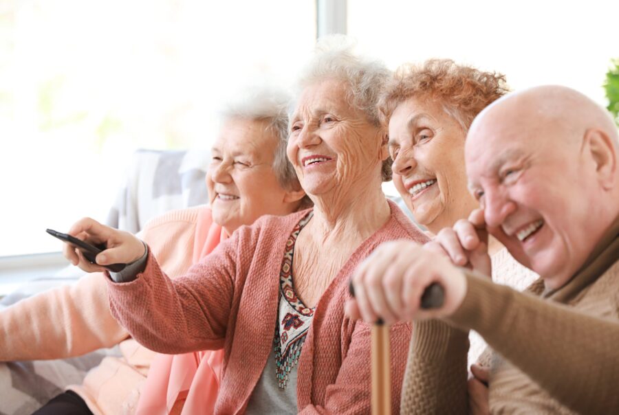 Groupe de 4 seniors assis sur le canapé avec des télécommandes dans les mains regardant la télé et souriant