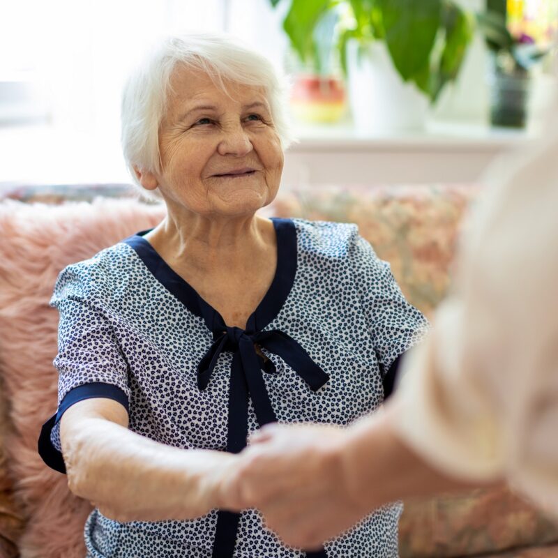 Femme senior assise dans un canapé avec une aidante qui lui tient les mains